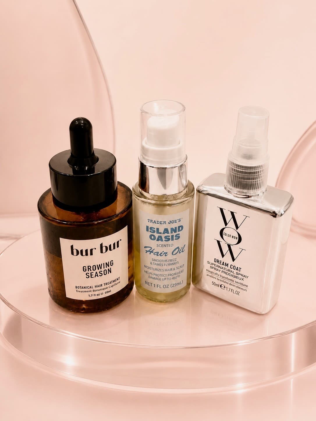 Three hair products photographed on a pale pink surface, including a treatment, an oil and a smoothing spray