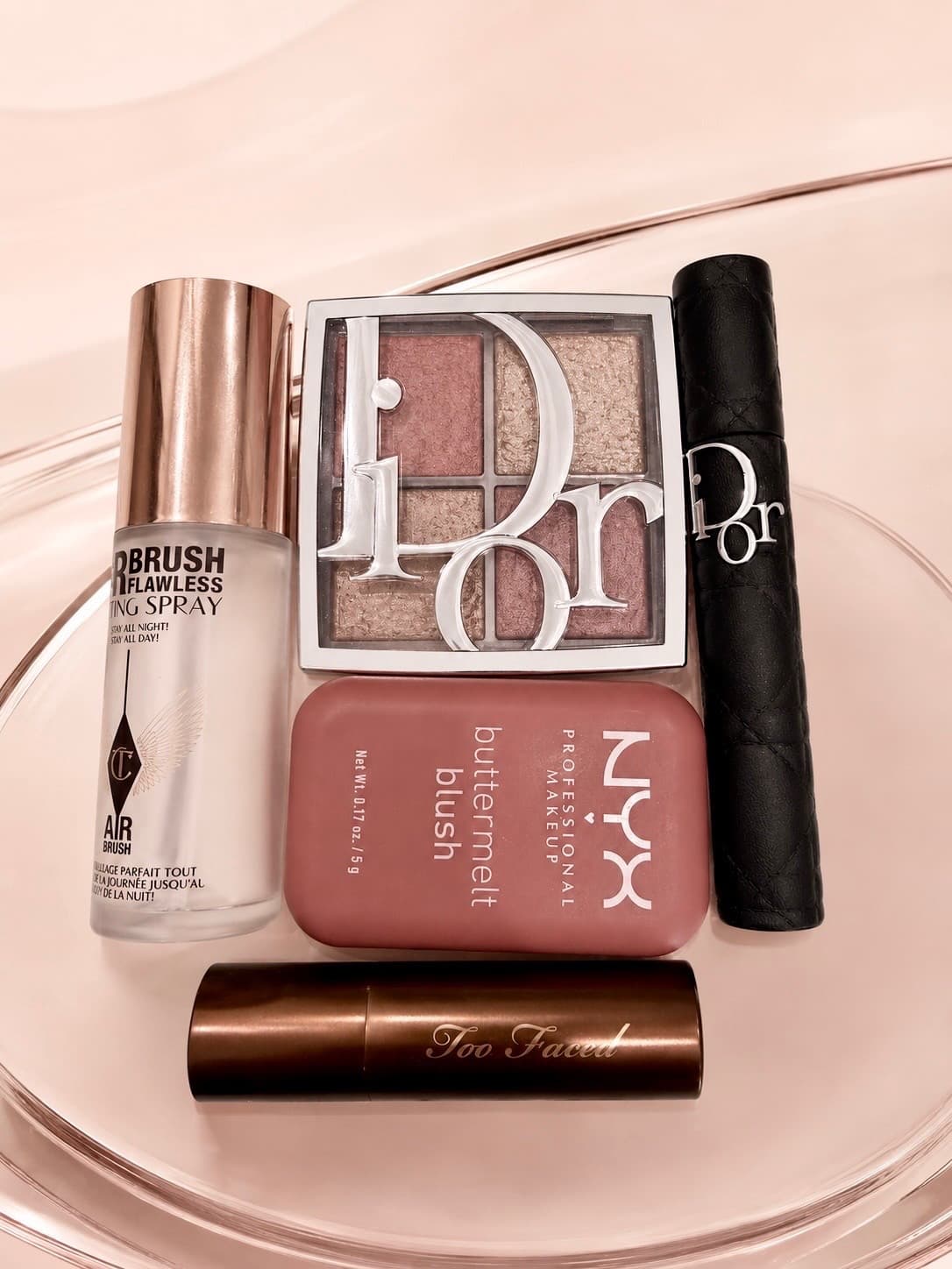 A flat lay of makeup including an eyeshadow palette, blush, mascara and setting spray